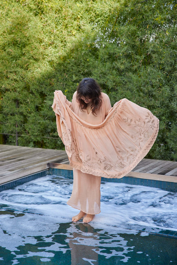 Artist Emilie Halpern stands in a peach dress that she lifts above her knees as she stands on the edge between her modern hot tub and pool.