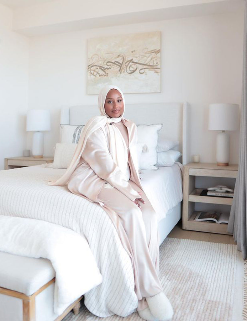 Influencer and blogger Aysha Harun sits on her ivory upholstered bed with neutral ivory and cream colored linens. Next to the bed are two matching square light wood nightstands with ivory cylindrical lamps.