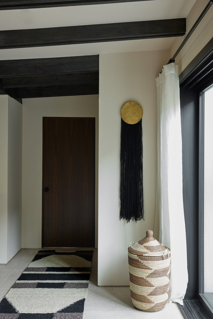 An Expedition Subsahara Astou brown and natural woven storage basket sits in a hallway under a black and gold wall hanging and next to a black, gray and ivory runner rug.