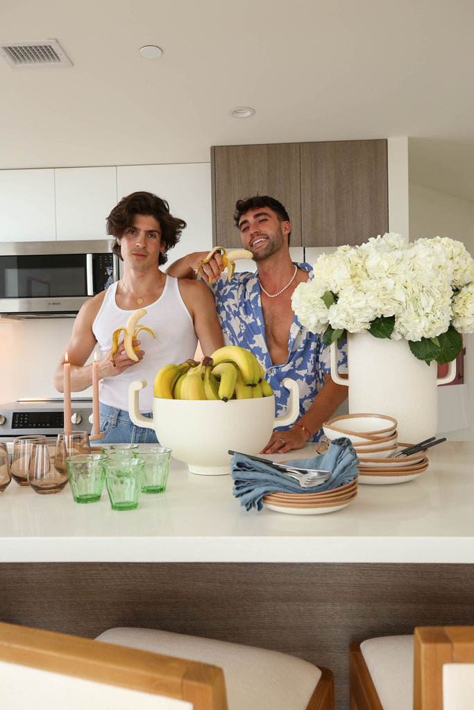 Chris Lin and Brock Williams of Yummertime stand in their modern LA kitchen, each holding a banana. A large white vase full of hydrangeas is on the right and a large white serving bowl full of bananas is in the middle.