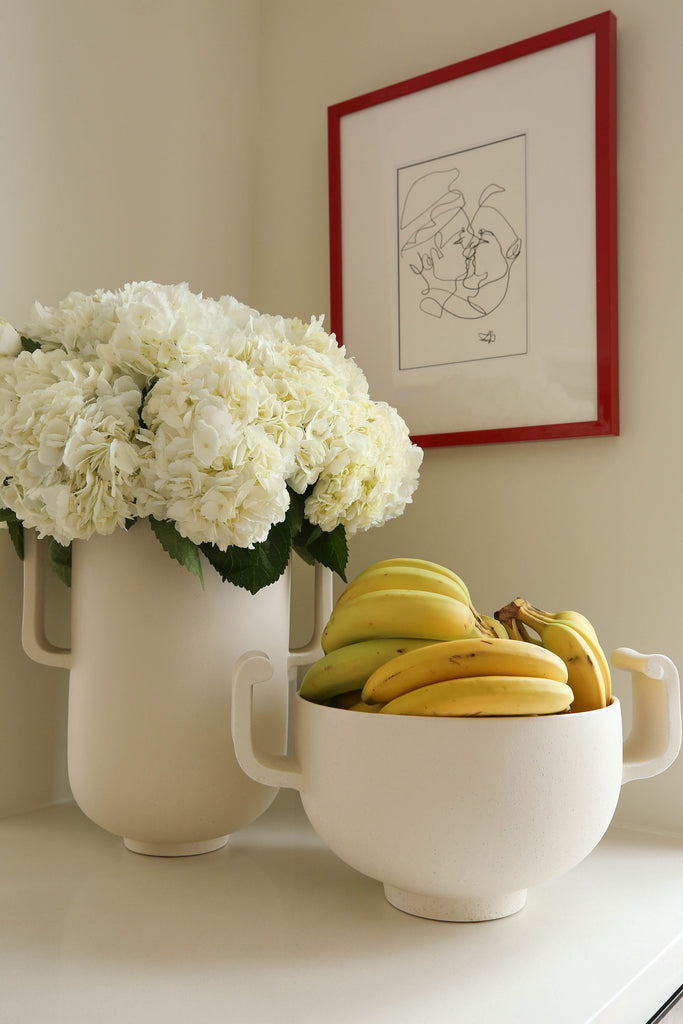 A tall ceramic white vase holds large white hydrangeas and sits next to the lower white ceramic Fallon serving bowl full of bunches of bananas.