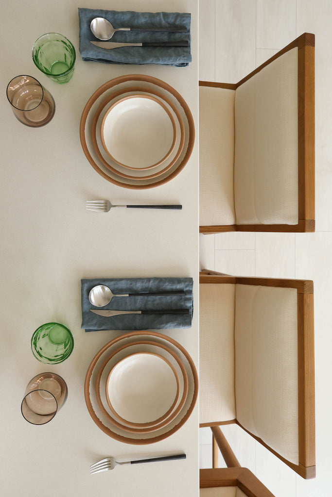 A down-shot shows two place settings with white glazed terracotta dinnerware, flatware with black handles, smoke Estelle stemless wine glasses, green water glasses, blue linen napkins, and two white upholstered Mirasol counter stools with wood trim.