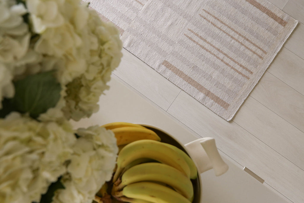 A down-shot with white hydrangeas, bunches of bananas in a white ceramic bowl, and a small, neutral striped flat rug.