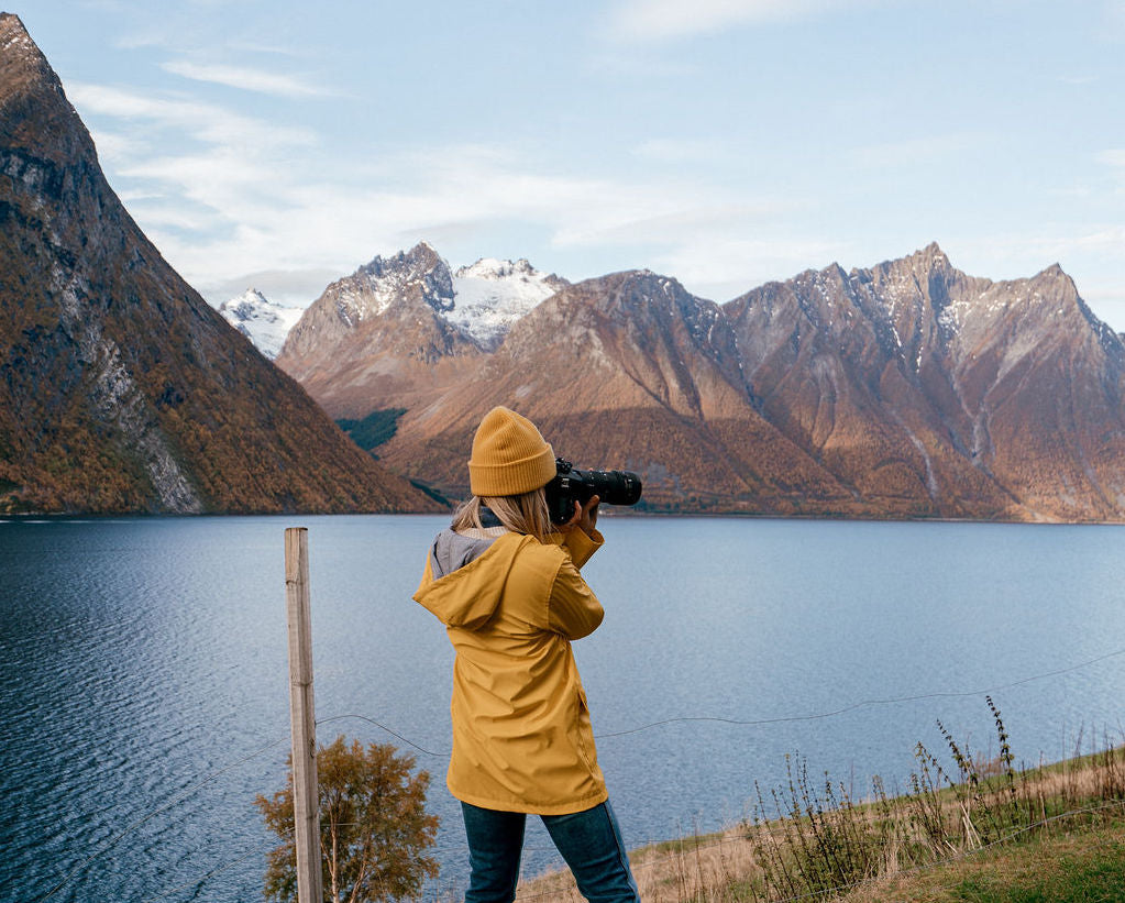 Photographer Carley Rudd takes a photo of mountains and a lake while standing in front of a wire fence.