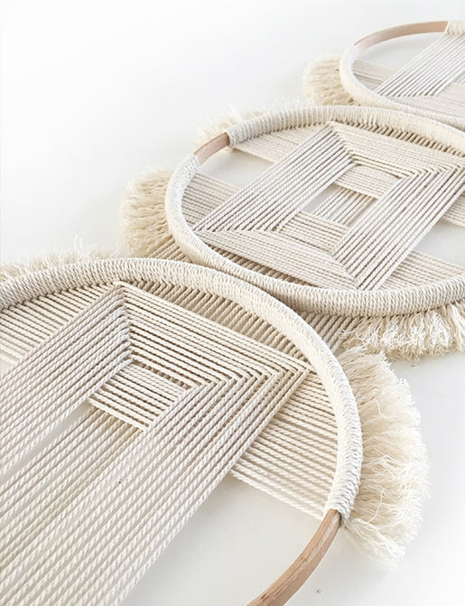 A long Studio Nom wall hanging with three concentric hoops with square-patterned weaving in the hoops, lays on a counter.