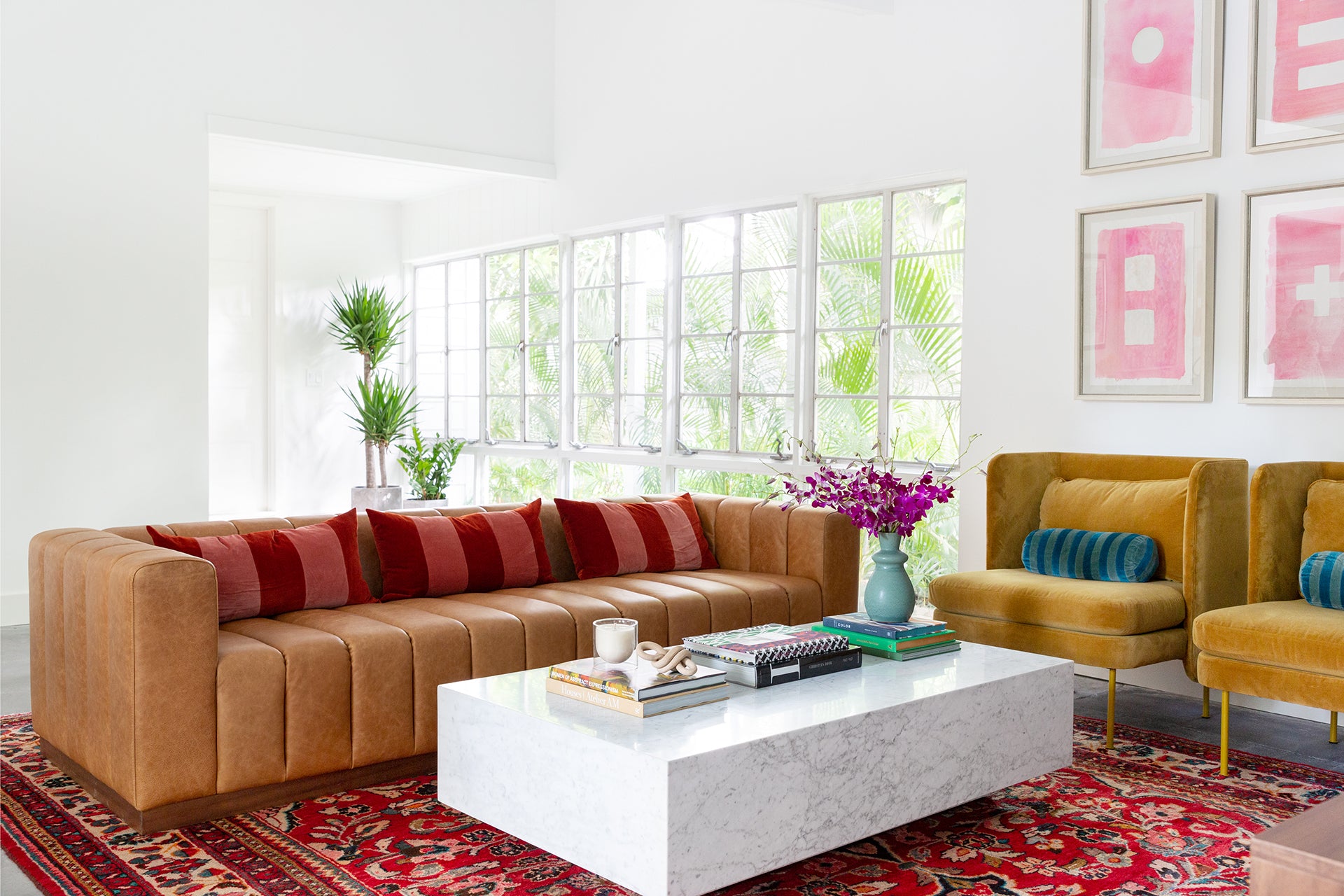 A camel brown squared sofa with red and coral striped throw pillows sits in a seating area with two gold velvet accent chairs with teal striped velvet pillows. A large white marble coffee table sits in the middle atop a traditional patterned red rug.