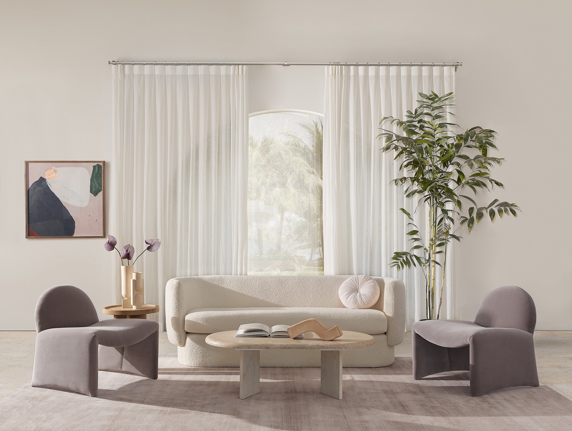 A light and airy living room has a curved Zaha boucle sofa with two velvet armless curved accent chairs, a light wood coffee table and long white curtain panels.