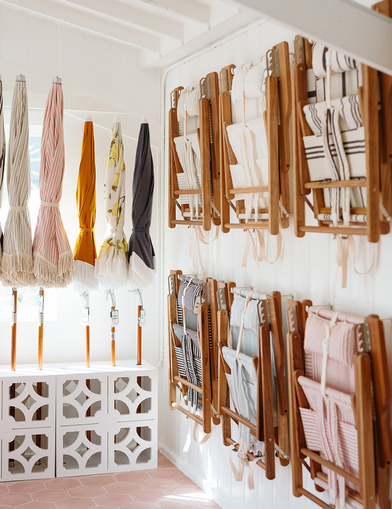 Closed Business & Pleasure Co. patio and beach umbrellas line a wall in a white holder. Two rows of their beach chairs with teak frames and woven seats are folded and hang on a wall.