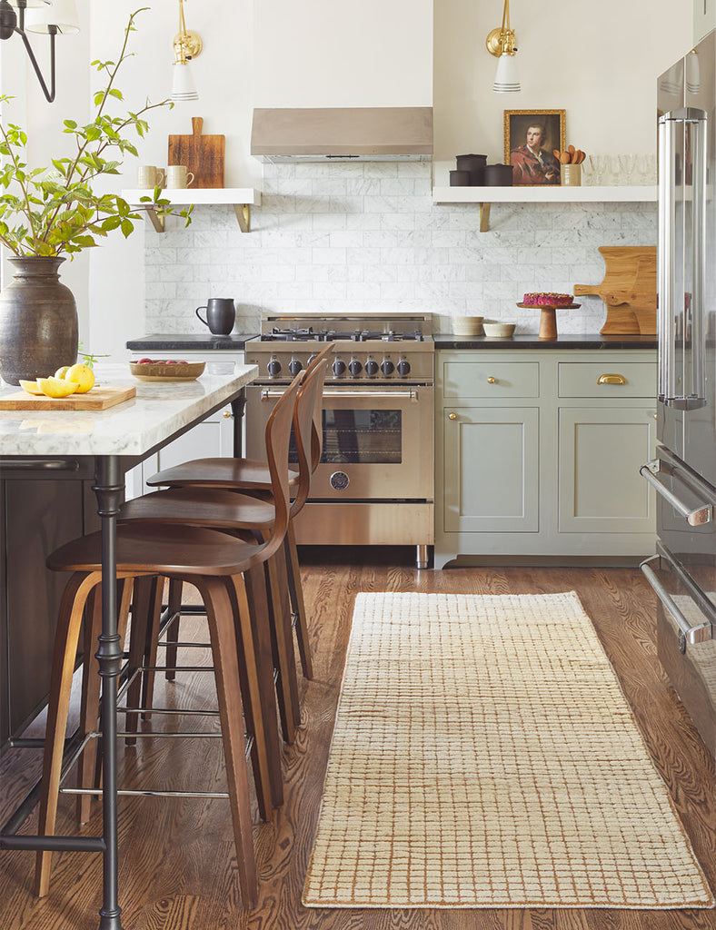 The brown and neutral subtle check Uma Runner Rug sits in a kitchen in between a refrigerator and a marble, wood and iron island flanked with medium brown wooden barstools.