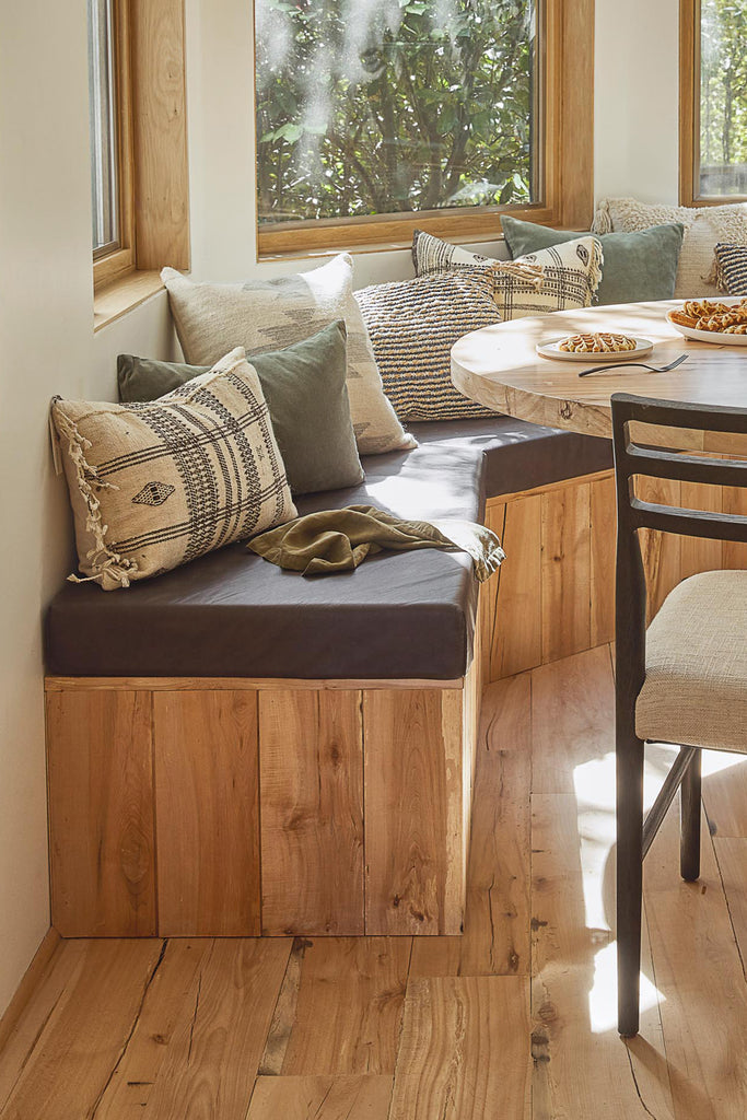 A built-in banquet covered in light wood paneling and topped with a thick gray cushion and neutral throw pillows offers seating to a round light wood dining table.