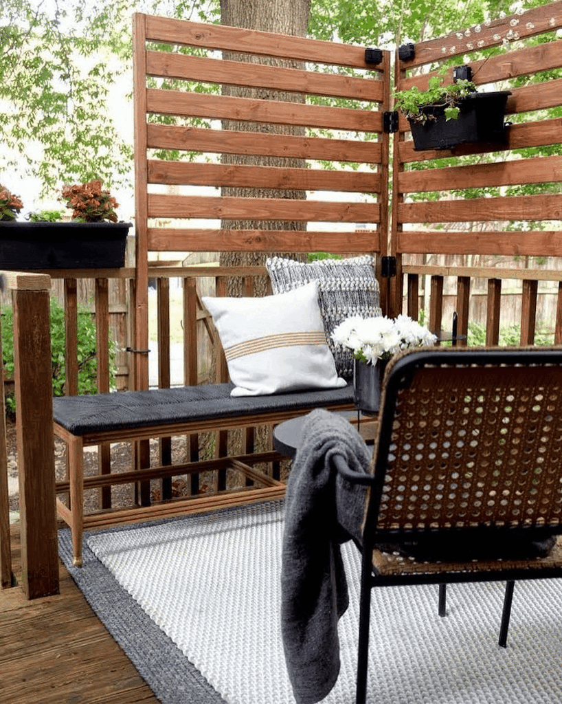 An seating area on a deck is enclosed with wooden slats and a built-in bench has a black woven seat and throw pillows. Designer Malcom Simmons arranges a gold throw pillow on a bed with mauve bedding and a paneled headboard.