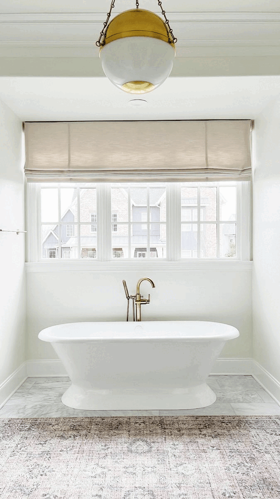 A free standing bathtub image flips to include a tall arched gold framed mirror, hanging white towels, a small round table with a decorative accessories on it and a small box holding white towels.