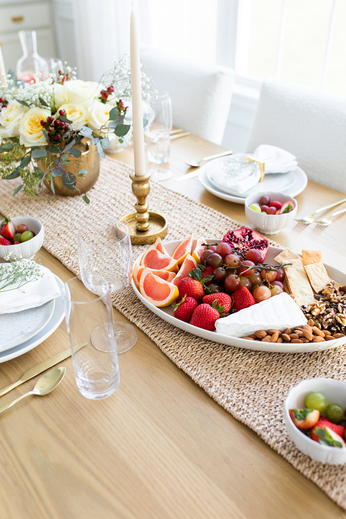 A white platter of nuts, cheeses, and fruits sits on a woven seagrass table runner on a light wood table.