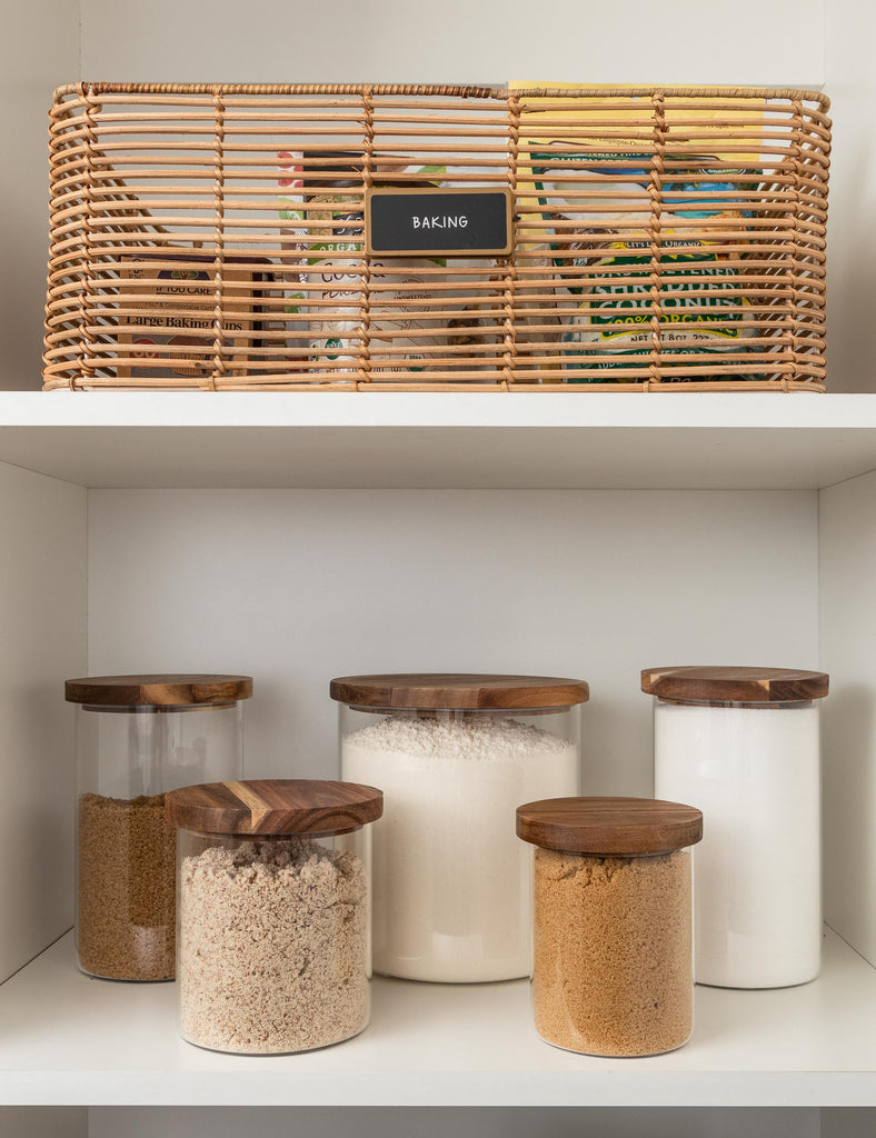A rattan basket sits on a shelf holding baking essentials above a shelf full of NEAT Method glass jars with wooden lids holding sugars, flour, and other baking dry goods.