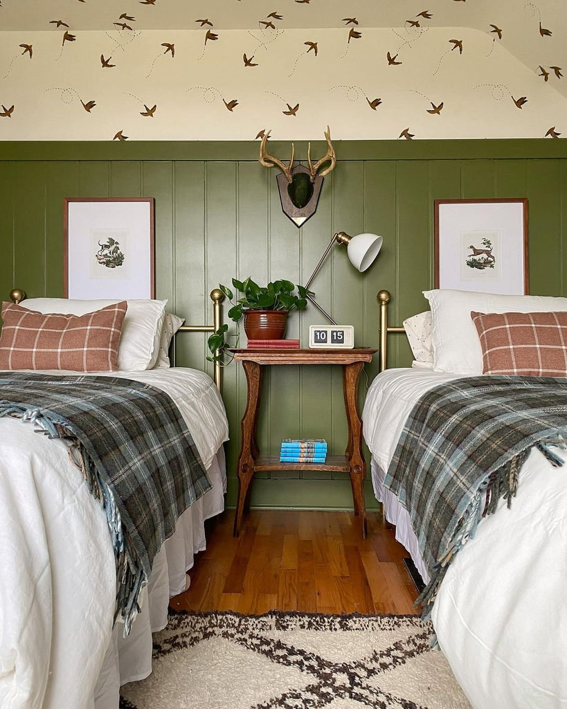 Two twin beds with ivory bedding with blue and gray plaid blankets have a small wooden nightstand in between them with a small plant and a small desk lamp. The wall behind the beds is has neutral wallpaper with birds above olive green wainscotting.
