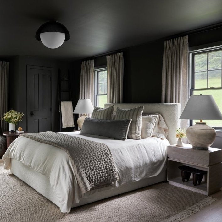 An ivory upholstered bed with ivory bedding and gray throw pillows and throw blanket contrast against dark gray walls. A light wood nightstand with a neutral ceramic table lamp sits on the side table.