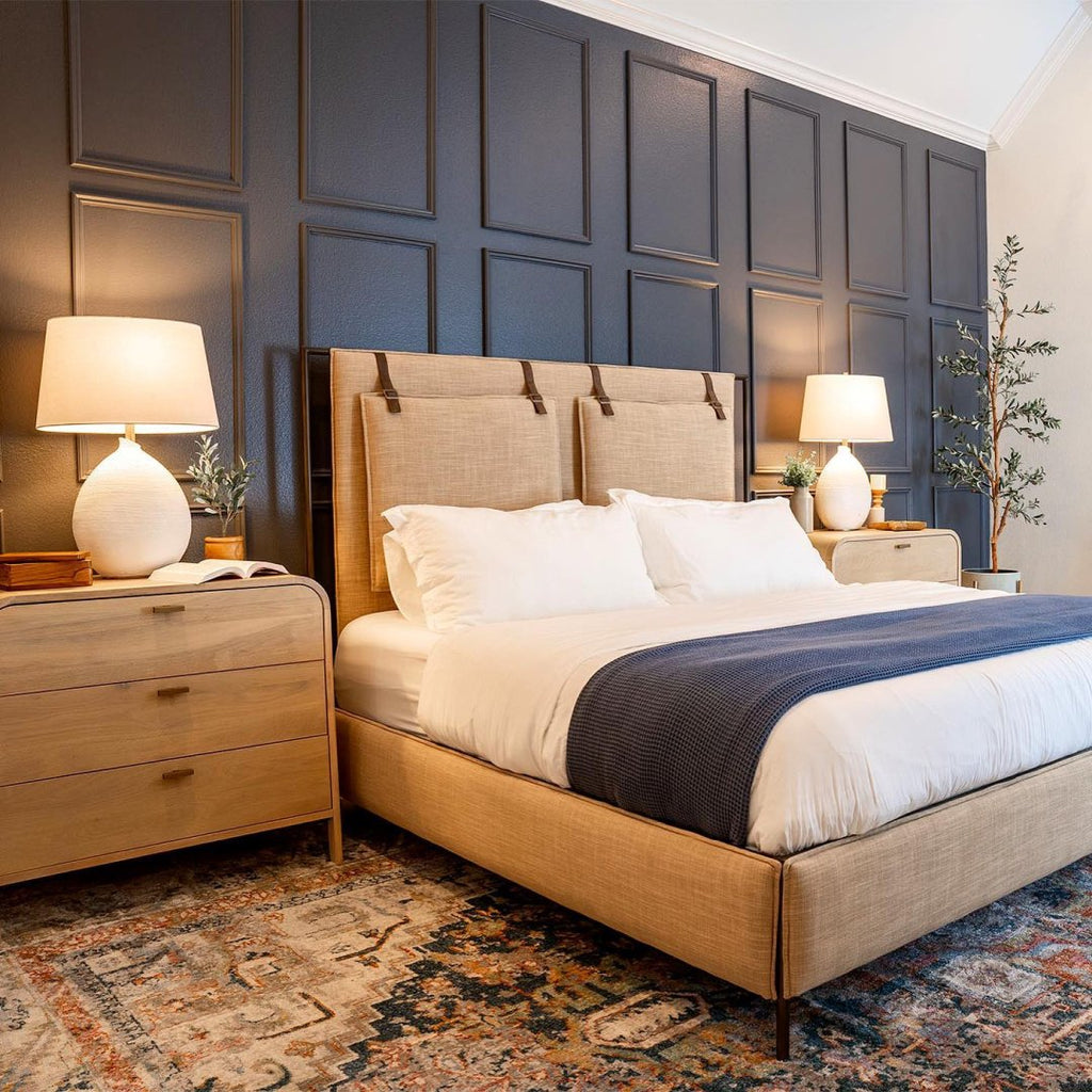 A peach-colored padded headboard has white bedding and a dark gray blanket on the end. Two light wood rounded nightstands flank the bed with white ceramic lamps. A dark paneled wall is behind the bed.