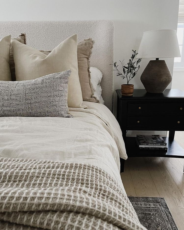A boucle headboard anchors this bed with neutral linen bedding and throw pillows. A dark wood nightstand is next to the bed with a small plant and a dark table lamp with white shade.