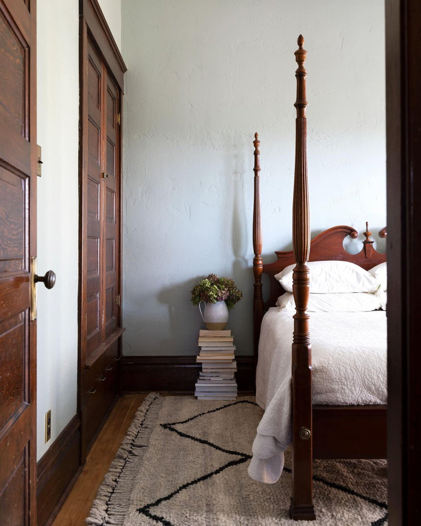 A traditional dark wood 4-poster bed has a white quilt on it. A stack of beds next to the bed has a pitcher on top with flowers in it. The modern Aya Moroccan Shag Rug grounds the bedroom.