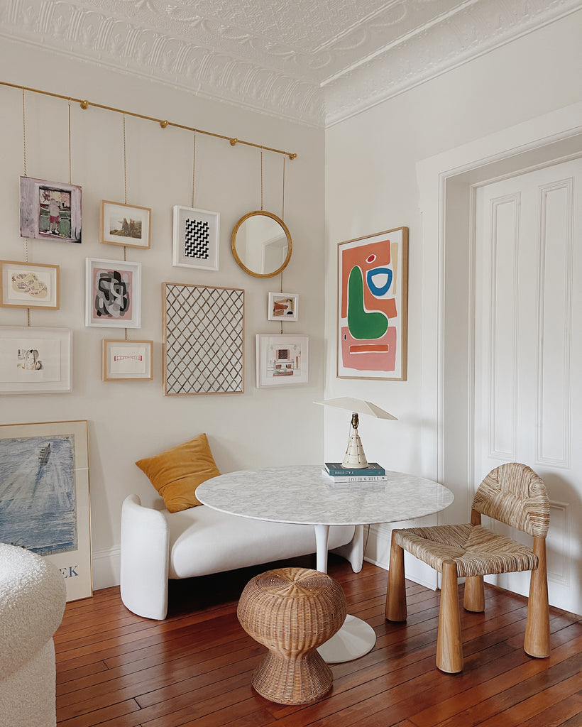 A round white dining table sits in a corner with a white bench and woven light wood Heidy chair. A small woven ottoman sits next to the chair and a gallery wall of colorful modern art hangs over the bench.