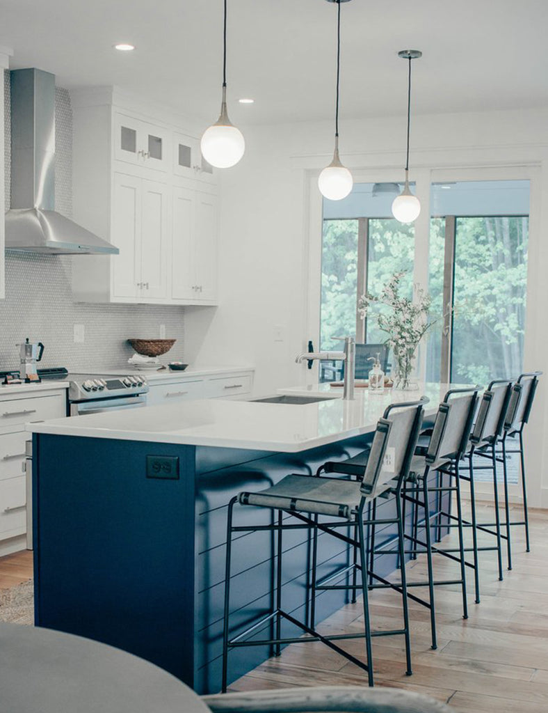 Three globe pendant lights hang over a long kitchen island with black cabinets and white quartz countertops. Four upholstered iron bar stools sit at the counter.