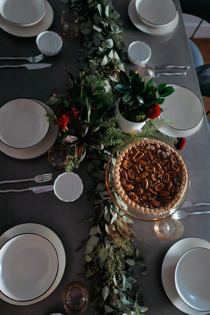 A pecan pie rests on a peach colored Estelle cake stand on a wooden table with green garland. White place settings and simple flatware is set on the table.