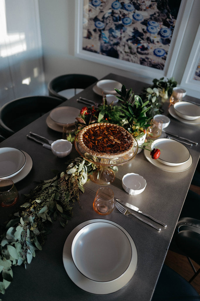 A dark wood table with green garland in the middle holds six white place settings with plates and bowls flanked by simple flatware. An Estelle peach colored glass cake stand holds a pecan pie.