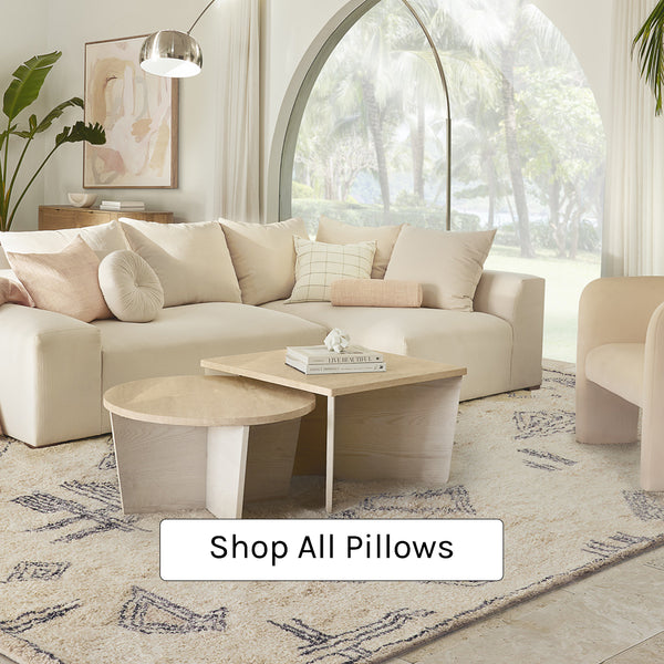 Cream and blush throw pillows sit on a modern cream colored sofa. A neutral plush area rug with black geometric shapes anchors the space and a small round and small square light wood table come together to create a coffee table in the center of the room. A large arched nickel floor lamp hangs over the sofa.