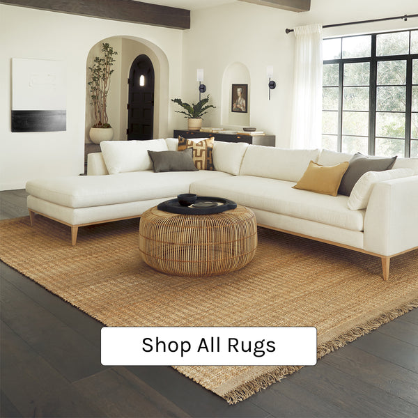 A modern ivory sofa with chaise sits on a jute rug with fringe. On the sofa are moss green and mustard throw pillows. A round rattan coffee table sits in the middle.