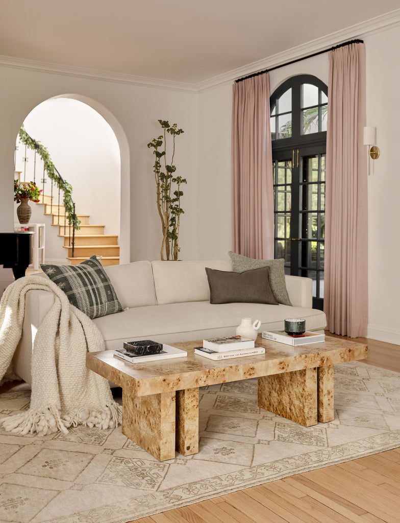 An ivory sofa 
 with black, brown and gray throw pillows sits in a vintage-inspired living room. Across from the sofa is a burl wood coffee table, and blush colored drapes frame a window.