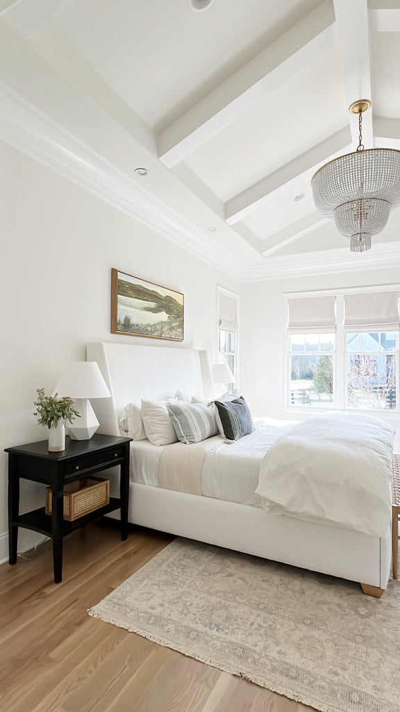 A classic bedroom design with a tall white upholstered bedframe with white and ivory bedding and gray and black striped throw pillows. A crystal chandelier hangs over the bed and a black nightstand with a white lamp, a small vase with flowers and a woven box sits next to the bed.