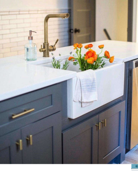 Farmhouse sink with blue vanity, antique brass euro cabinet pulls and an antique brass faucet. The sink has a towel and orange flowers in the basin. 