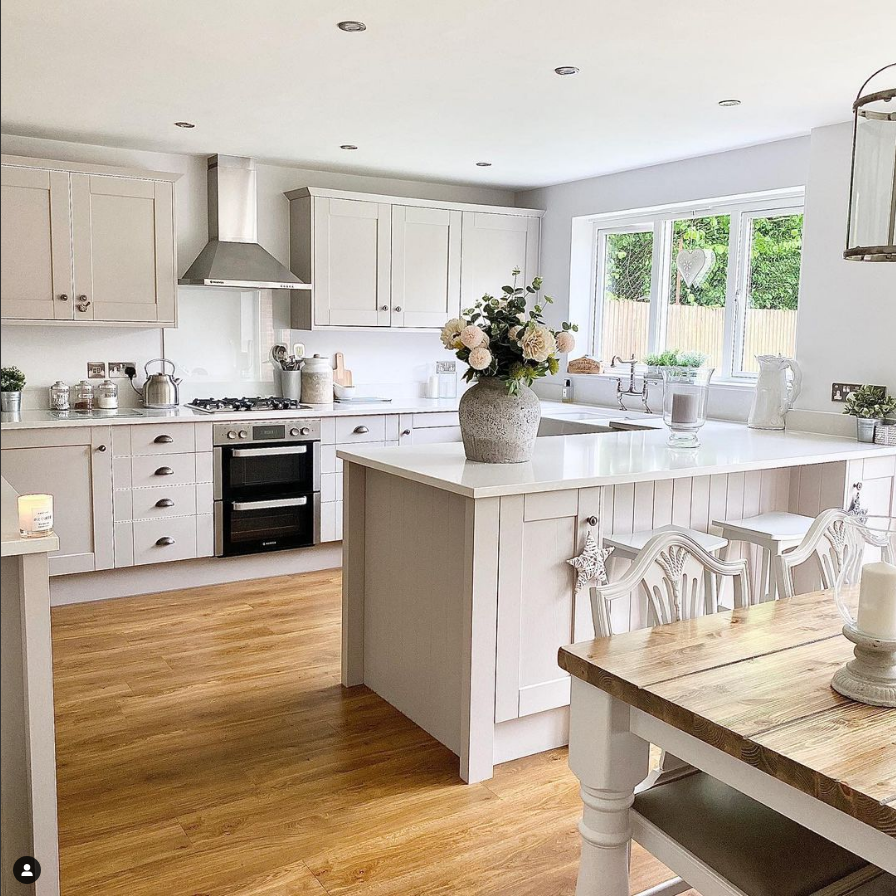 Large modern country kitchen with warm hardwood floors and white cabinets. The kitchen cabinets have weathered nickel hardware and black appliances. 