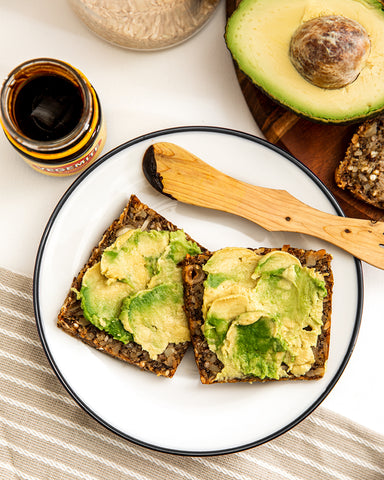 Flat lay of two pieces of seed bread with avocado and vegemite. Jar of vegemite and half an avocado in the background