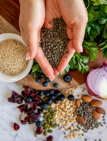 Lady holding lentils in hands above a table scattered with a bunch of whole foods including nuts, seeds, legumes