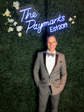 Jason Mitchell Kahn standing in front of a green backdrop with blue lit sign wearing a grey tuxedo.
