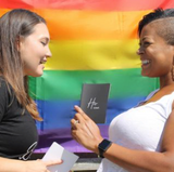 White woman (L) gazing at black womxn (R) while both holding vow books with a rainbow flag background