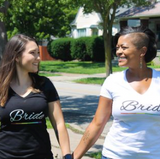 A white woman (L) holding the hand of a black womxn (R) - both wearing Bride shirts with rainbow line