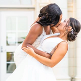 Black womxn (L)dipping and kissing white womxn (R) - both in wedding dresses