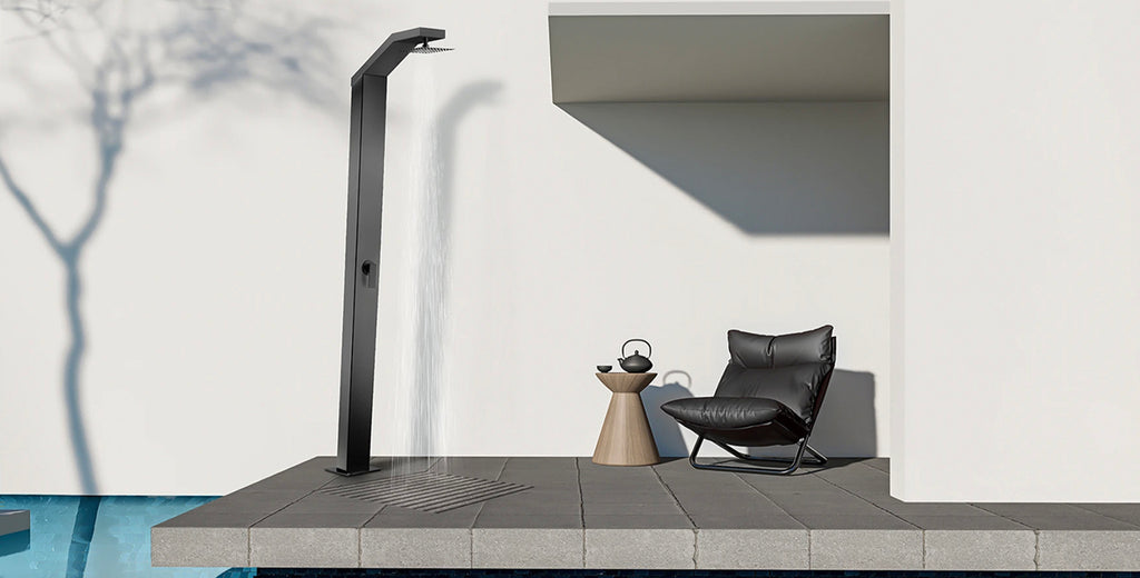 Columna de ducha independiente de exterior en acabado negro mate junto a una silla y mesa de exteriores, frente a una alberca en un día soleado