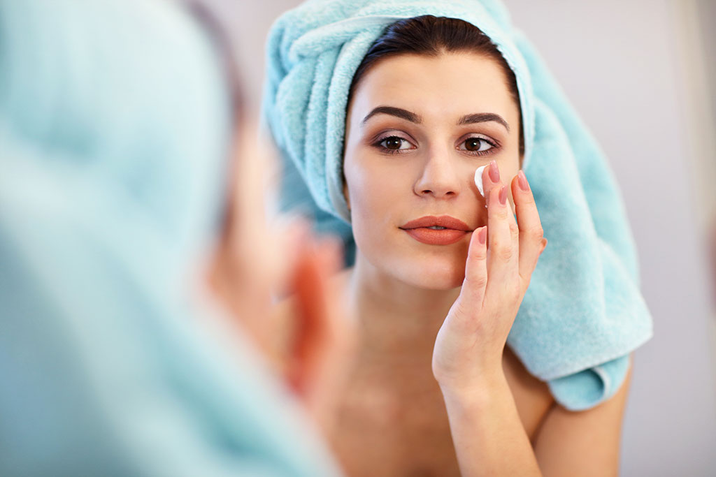 Young woman standing in bathroom and applying face cream in the morning