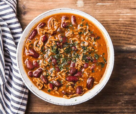 a neutral colored bowl full of chili on a wooden table with a blue and white striped napkin beside it.