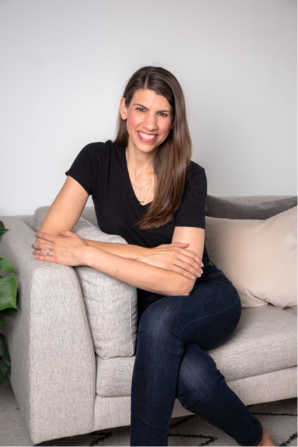 Jamie smiling sitting on the end of a grey couch with her legs and arms crossed