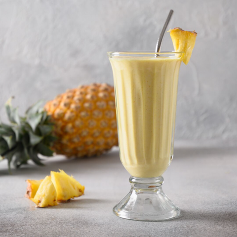 a pineapple lassi in a tall smoothie glass with a straw and a slice of pineapple on the side and a full pineapple lying behind it.