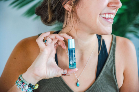 a woman with a green tank top holding the throat chakra roller blend between her first finger and thumb at the neck level smiling