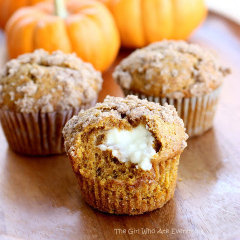 3 pumpkin cream cheese muffins with the cream cheese filling showing in the first muffin with a bite missing. Pumpkins are in the background of the muffins.