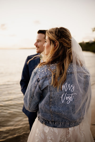 Bride and Groom standing next to the water with the bride wearing a personalized denim jacket over her wedding dress.