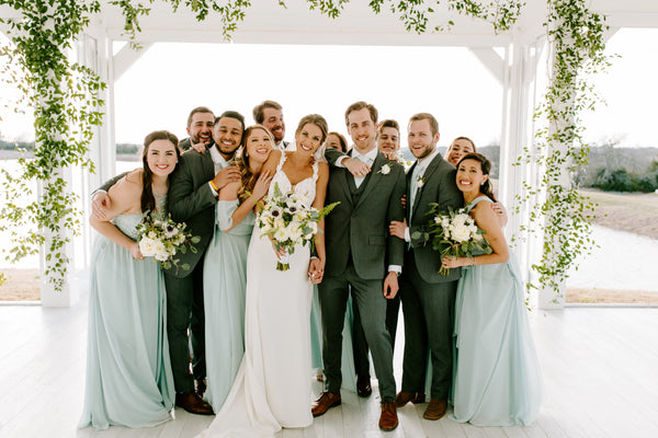 Bridal Party gathered together under a white pergola in greenery. Bridesmaids are wearing Kennedy Blue dresses in 'Slate Blue'.