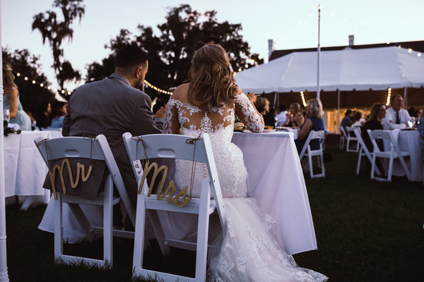 Bride and Groom sitting on wooden folding chairs with "Mr" and "Mrs" signs at their outside reception.