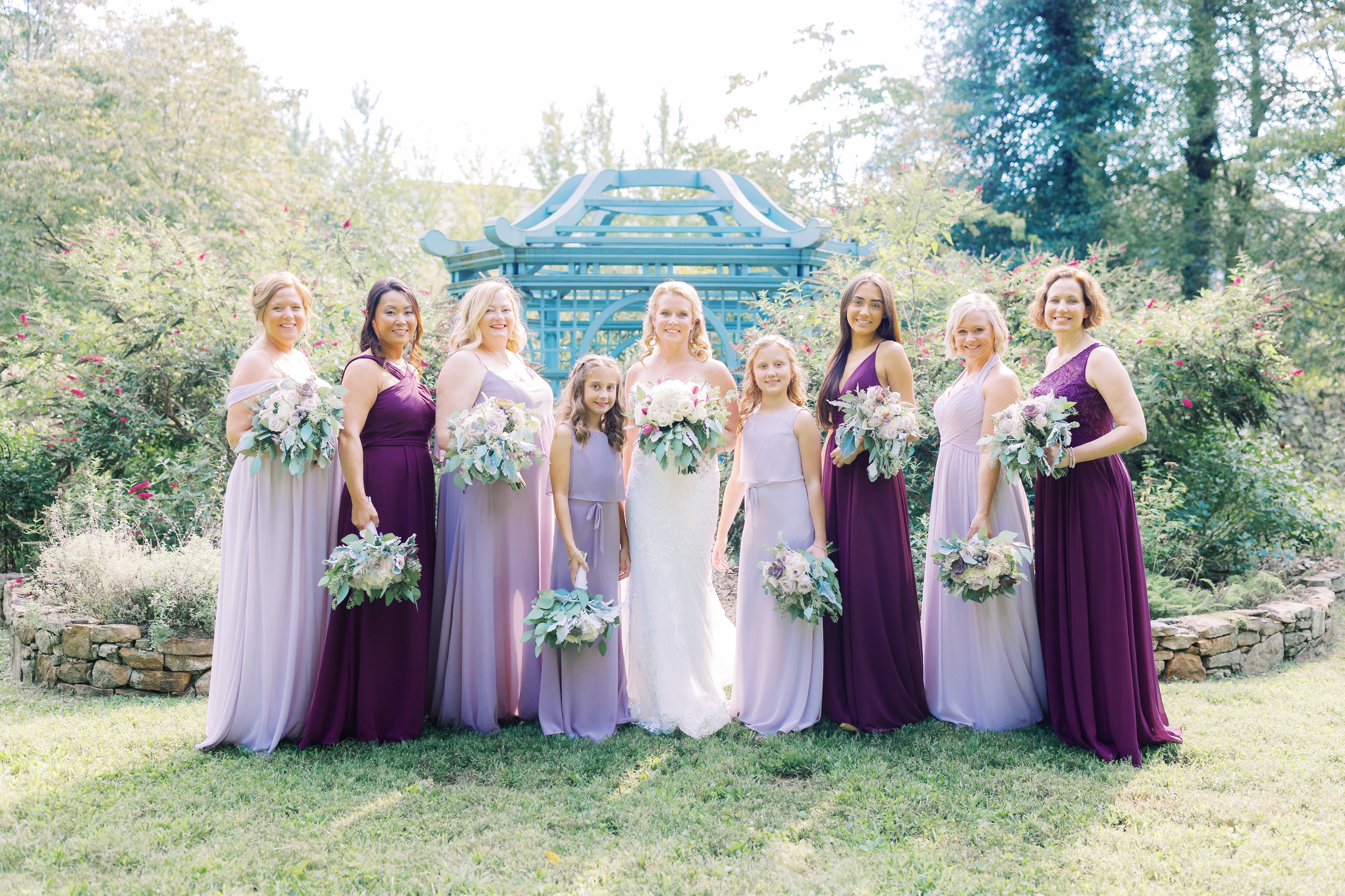 Bride and Bridal Party wearing Kennedy Blue Bridesmaid Dresses in French Lilac, Wisteria, and Eggplant.